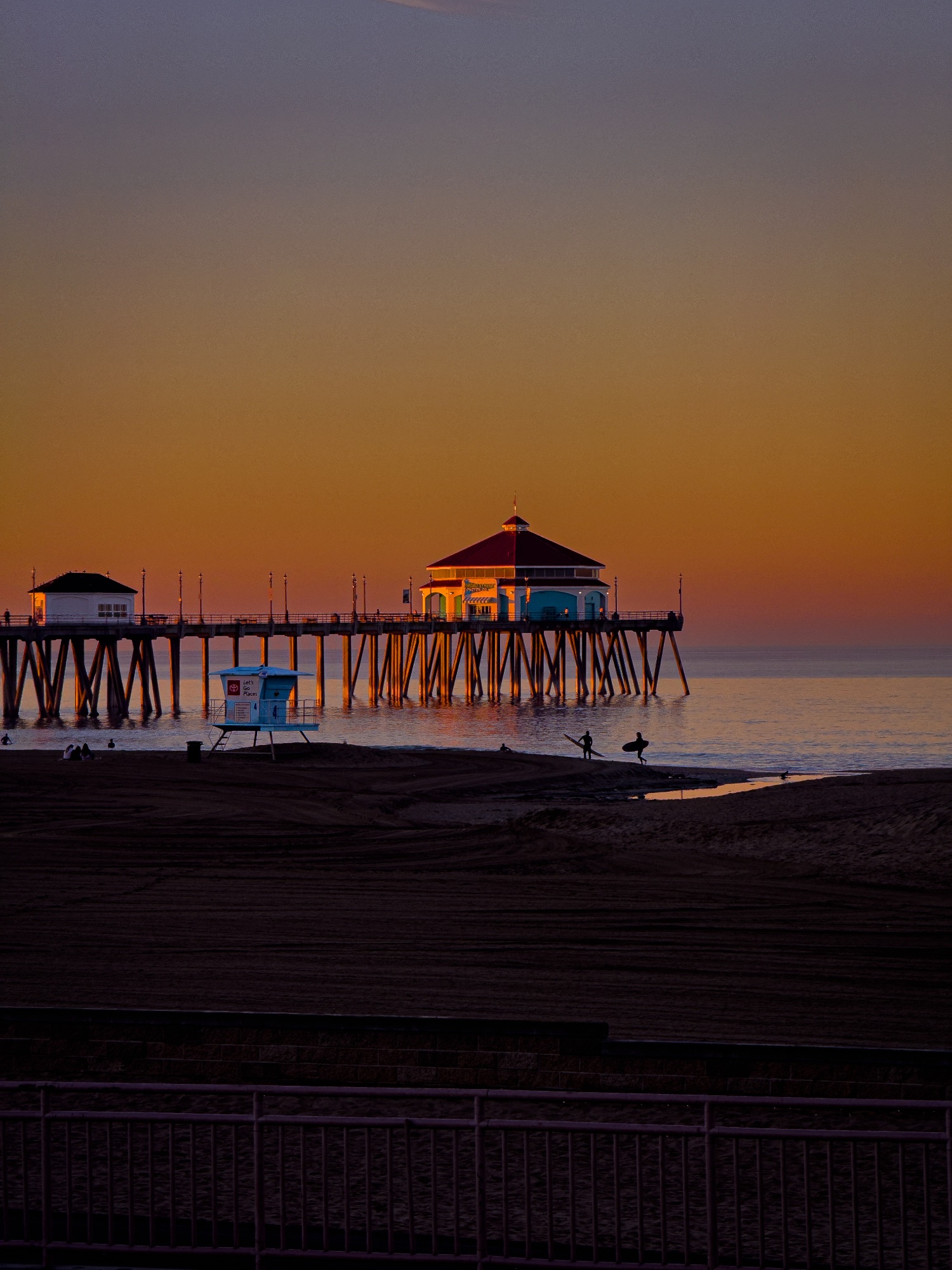 Sunrise view of the Huntington Beach pier with surfers and a lifeguard tower in the foreground